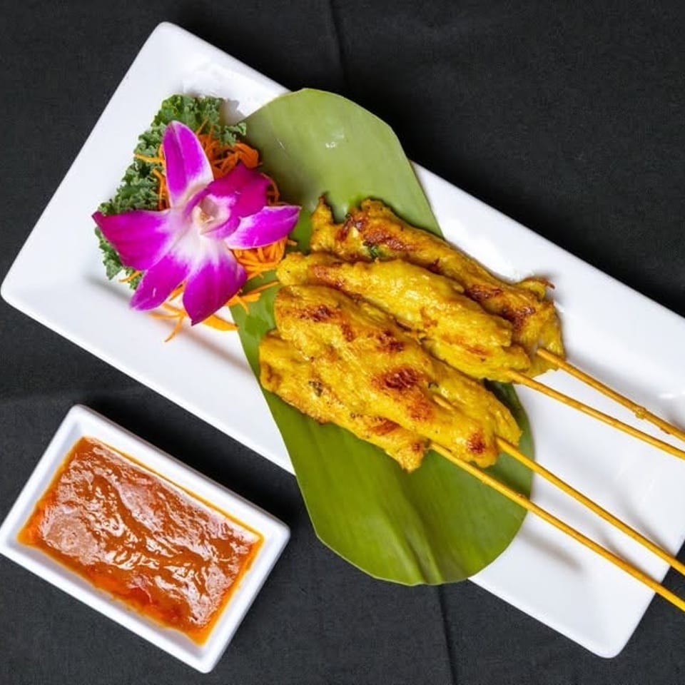 Chicken satay skewers on a banana leaf, with a purple orchid and dipping sauce.