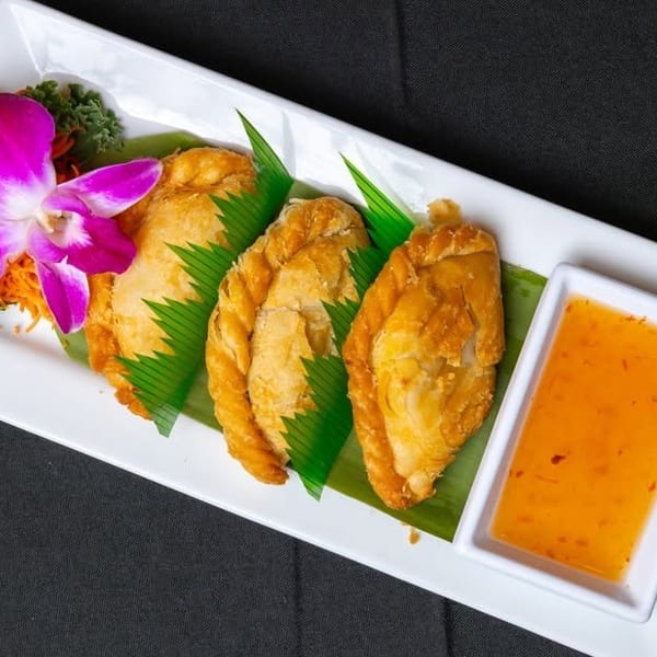 Three golden-brown curry puffs with sweet chili sauce and an orchid garnish on a white plate.