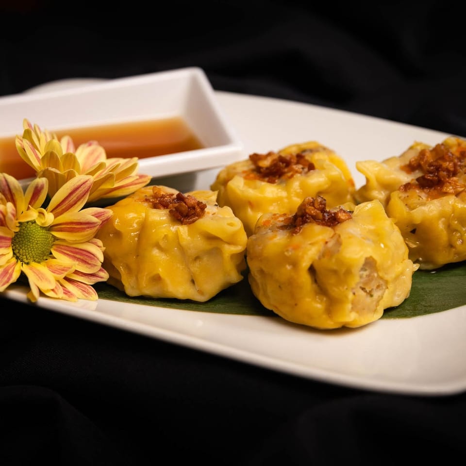 Four yellow dumplings with crispy toppings, a side of sauce, and a yellow flower on a white plate.