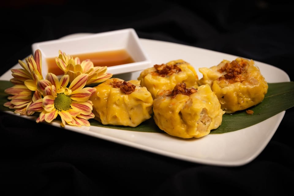 Four yellow dumplings with crispy toppings, a side of sauce, and a yellow flower on a white plate.