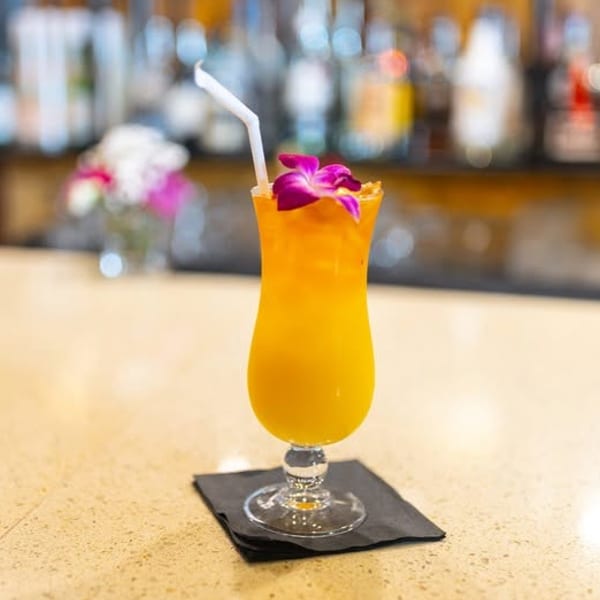 A vibrant orange cocktail with a purple orchid garnish and straw sits on a bar.