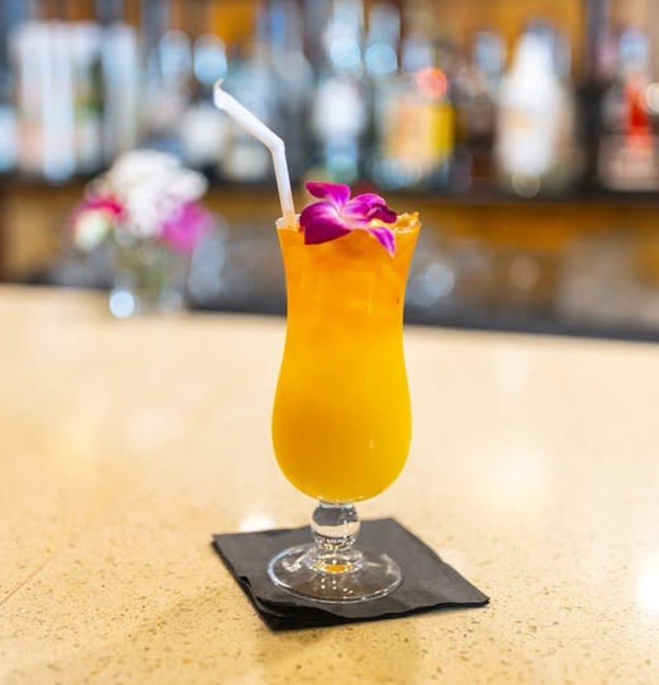 A vibrant orange cocktail with a purple orchid garnish and straw sits on a bar.