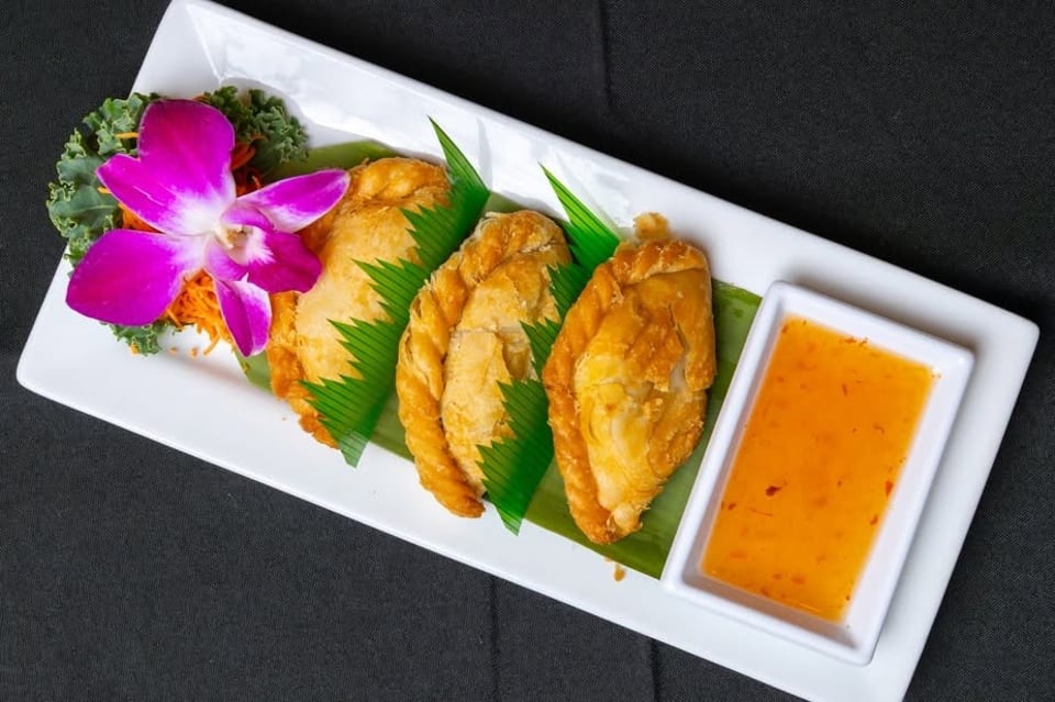 Three golden-brown curry puffs on a white plate with sweet chili sauce and an orchid garnish.
