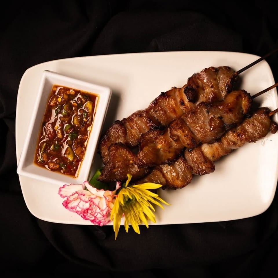 Grilled pork skewers with a spicy dipping sauce and floral garnish on a white plate.