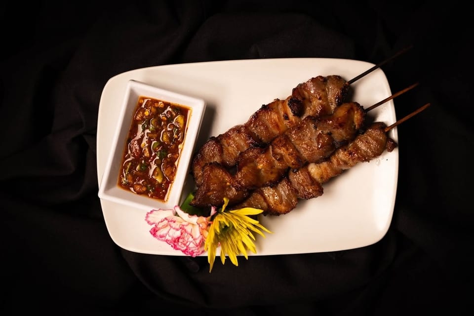 Grilled pork skewers with a spicy dipping sauce and floral garnish on a white plate.