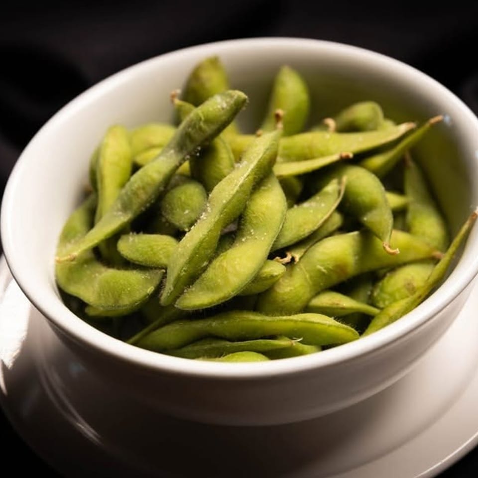 A white bowl filled with vibrant green, salted edamame pods on a dark background.