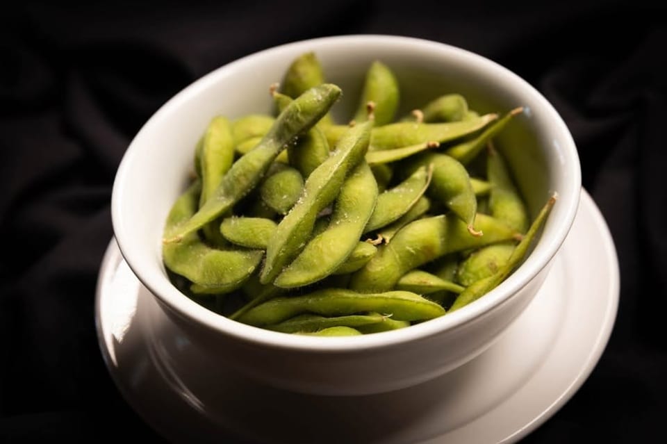 A white bowl filled with vibrant green, salted edamame pods on a dark background.