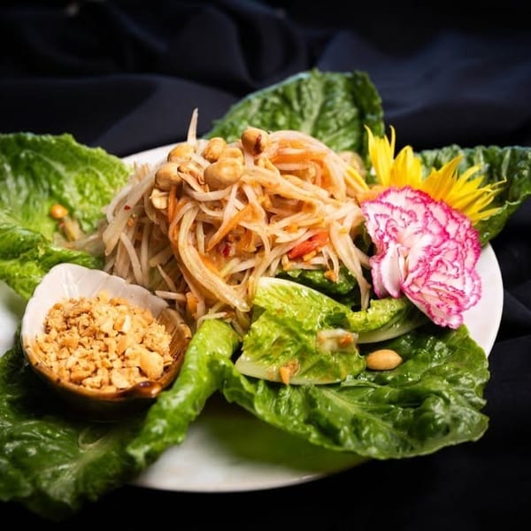 Papaya salad with peanuts, lettuce, and decorative flowers on a white plate against a dark background.