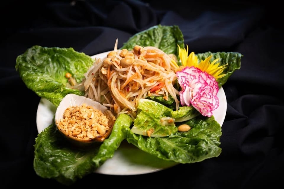 Papaya salad with peanuts, lettuce, and decorative flowers on a white plate against a dark background.