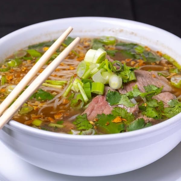 A white bowl of pho with beef, noodles, broth, green onions, and cilantro, with chopsticks.
