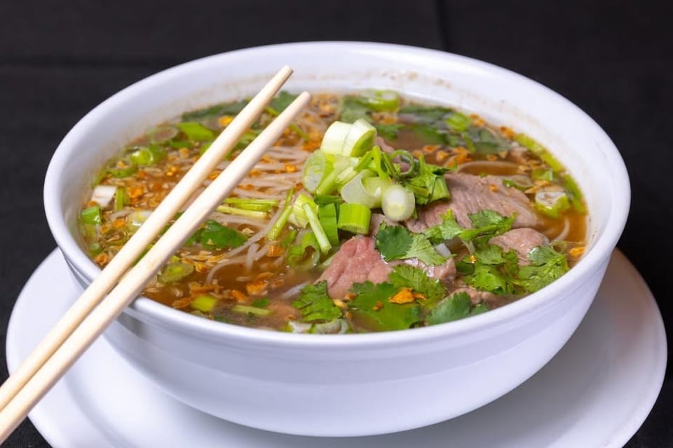 A white bowl of pho with beef, noodles, broth, green onions, and cilantro, with chopsticks.
